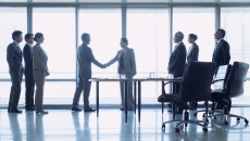 People standing in front of a large window with two of them shaking hands and six others standing and watching People standing in front of a large window with two of them shaking hands and six others standing and watching