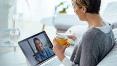 Person sitting on a couch holding a coffee cup while talking to a telehealth provider on their computer Person sitting on a couch holding a coffee cup while talking to a telehealth provider on their computer