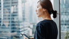 Person standing up while holding a phone and looking outs a window Person standing up while holding a phone and looking out a window