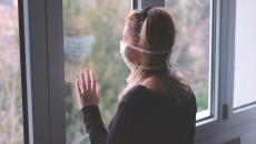 Person with a mask on standing by a window looking out Person with a mask on standing by a window looking out