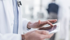 The body of a healthcare provider using a tablet while wearing a lab coat and stethoscope around their neck The body of a healthcare provider using a tablet while wearing a lab coat and stethoscope around their neck
