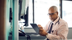 "Healthcare professional sitting at a desk with a window behind them while wearing a stethoscope around their neck and looking at a tablet" "Healthcare professional sitting at a desk with a window behind them while wearing a stethoscope around their neck and looking at a tablet"