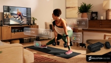 A woman lifting weights using the Peloton Guide while watching an instructor on her TV A woman lifting weights using the Peloton Guide while watching an instructor on her TV