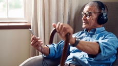 An older man in his home wearing headphones and using a smartphone. An older man in his home wearing headphones and using a smartphone.