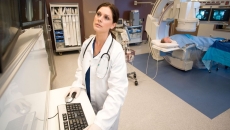 A medical professional looks at images while a patient receives a CT scan in the background A medical professional looks at images while a patient receives a CT scan in the background