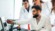 Three people wearing lab coats with one seated and pointing at screen Three people wearing lab coats with one seated and pointing at screen